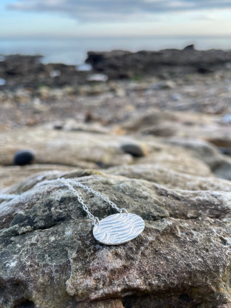 Silver Ripple Textured Oval Necklace on rocks by the sea, Kate Wimbush Jewellery
